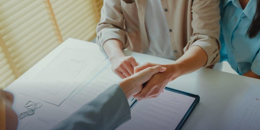 Two people shake hands over documents, a tablet, and keys on a table.